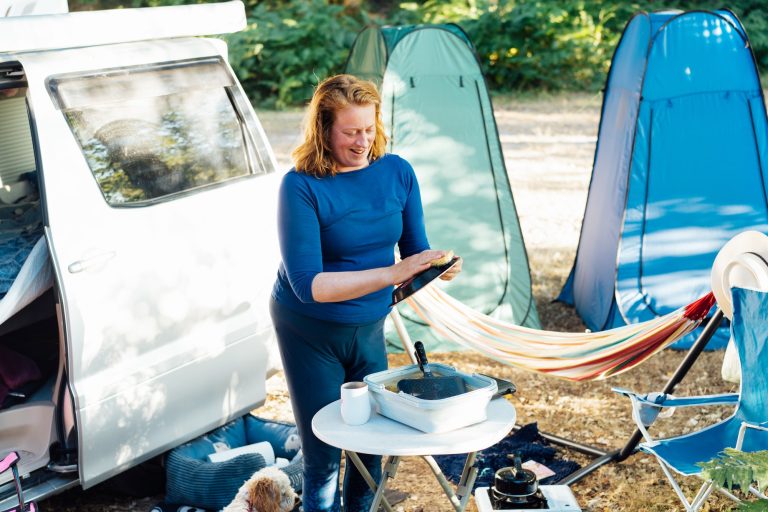 Middle age nomad woman washes dishes outside her camper van in forest camp during road trip.